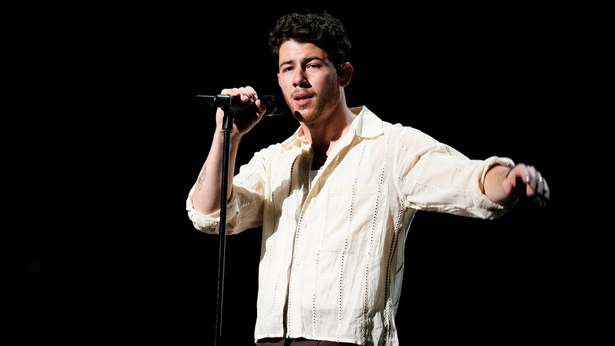 Nick Jonas stretches his arm on stage and holds the microphone with his other hand in a bright yellow shirt