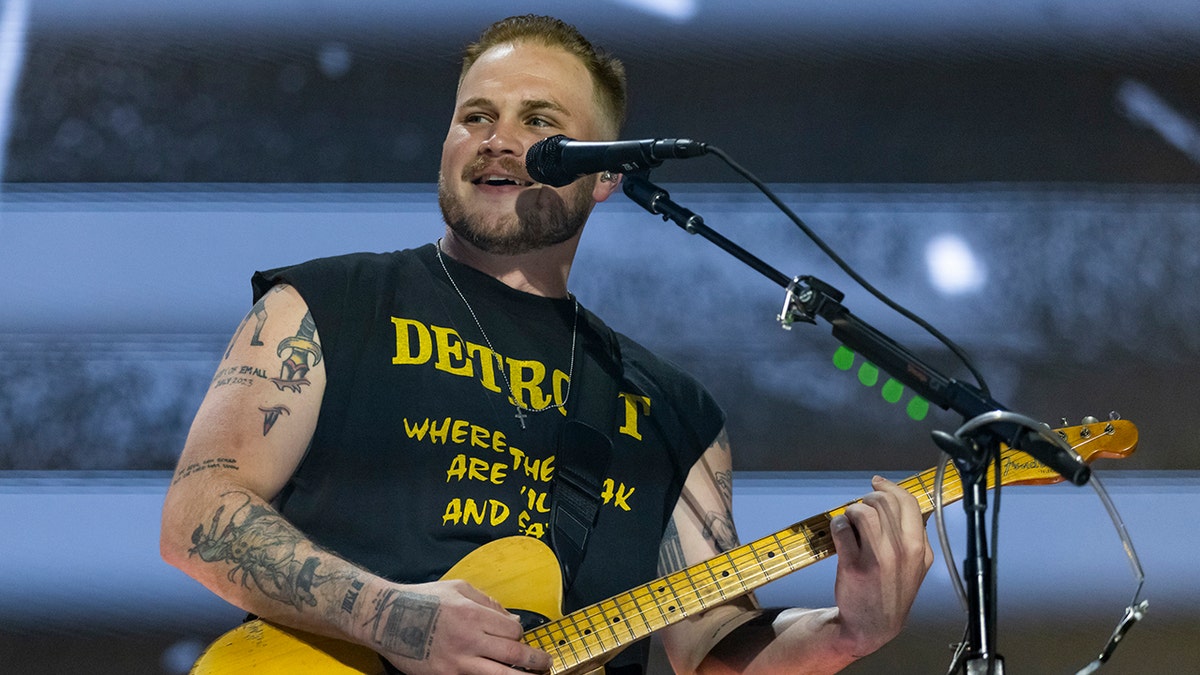 Zach Bryan on stage in a black cropped tank top with yellow lettering and a yellow guitar