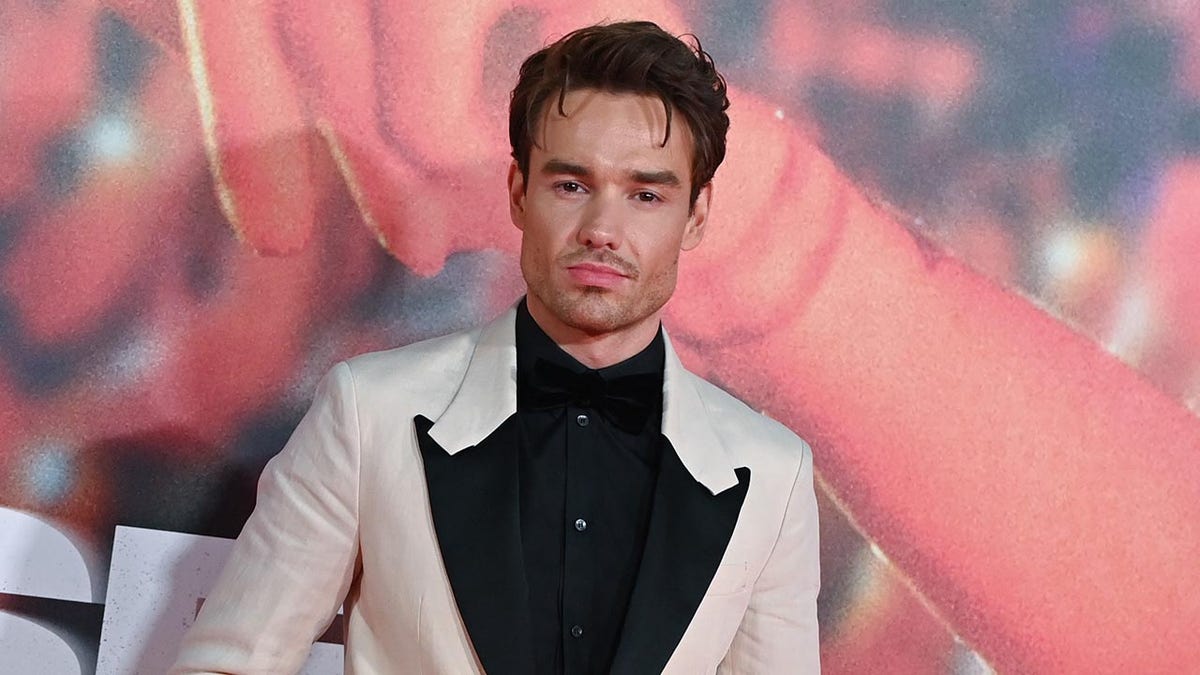 Liam Payne in a white jacket with black labels and a black shirt stands on the carpet in London