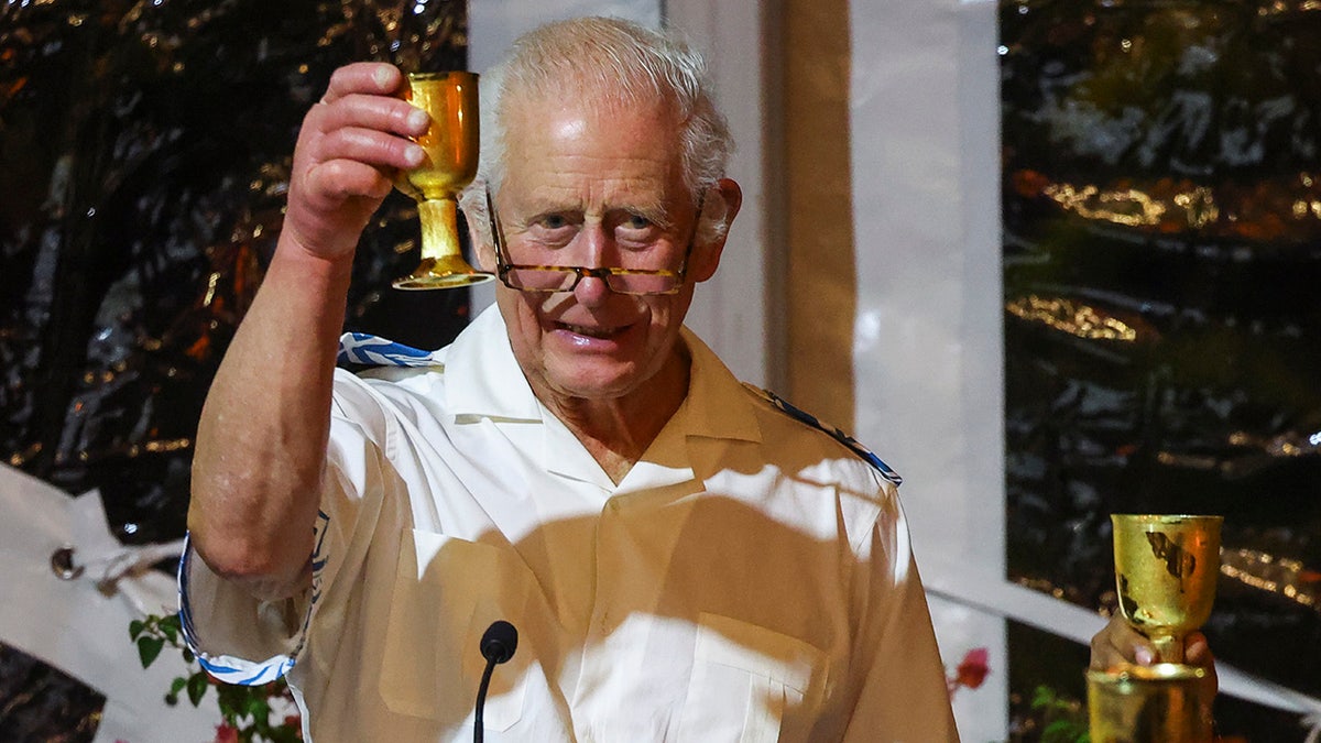 King Charles wearing a white shirt and glasses raising a gold glass.