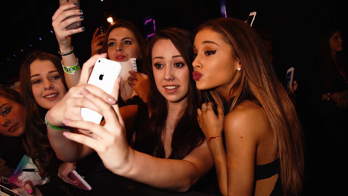 Ariana Grande pone cara de beso mientras se hace una foto con una fan que alarga los brazos para hacerse un selfie con su iPhone