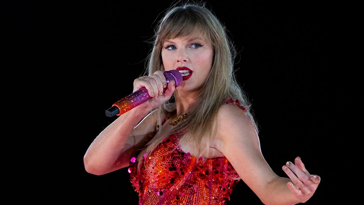 Taylor Swift sings into a shimmering orange-purple microphone and looks to the left of the stage in a red-orange bodysuit