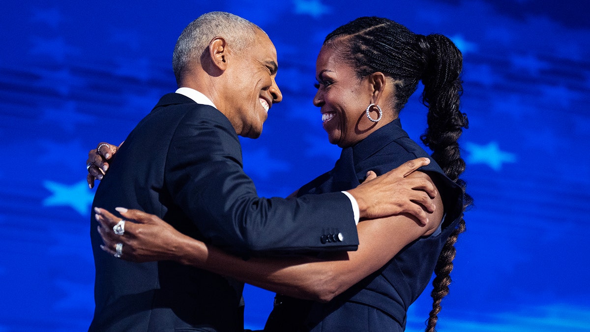 Barack Obama sorri ao cumprimentar a esposa Michelle Obama em um vestido azul marinho no palco e eles se abraçam