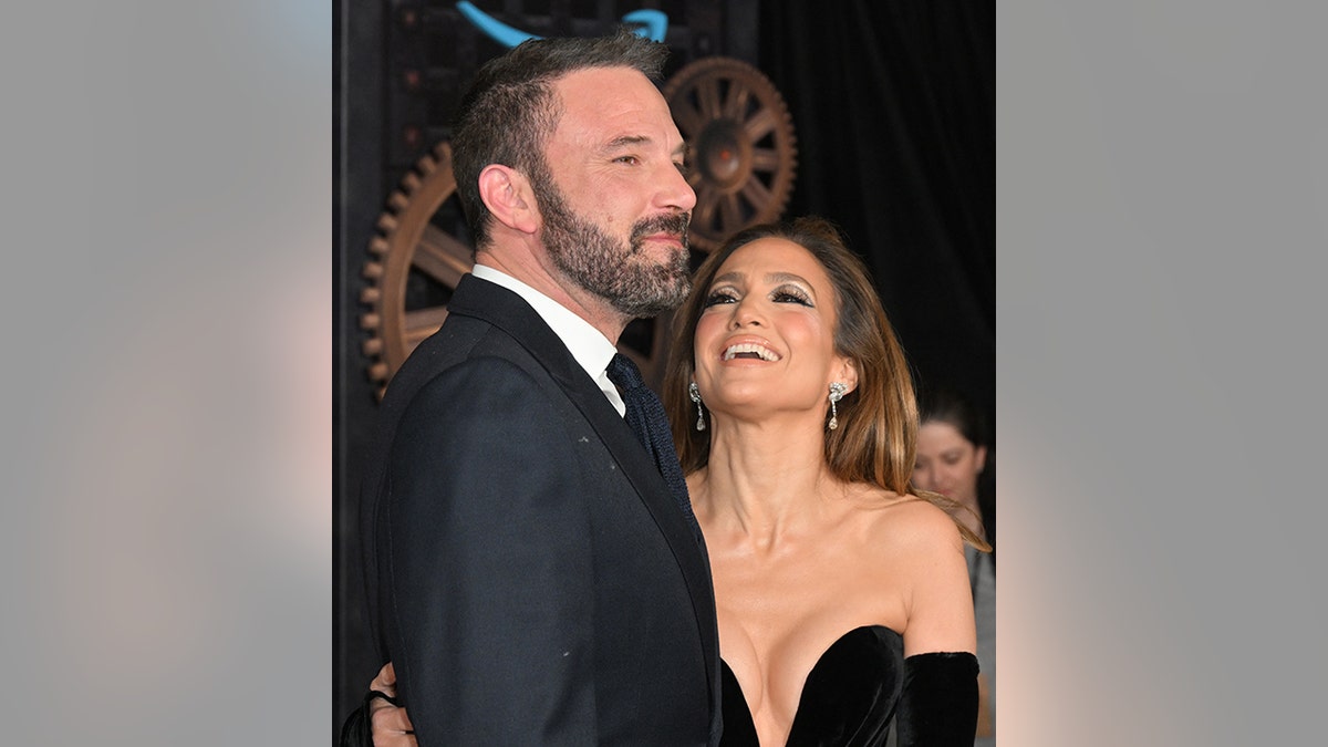 Jennifer Lopez em um vestido preto sem alças olhando para cima e rindo de Ben Affleck de terno