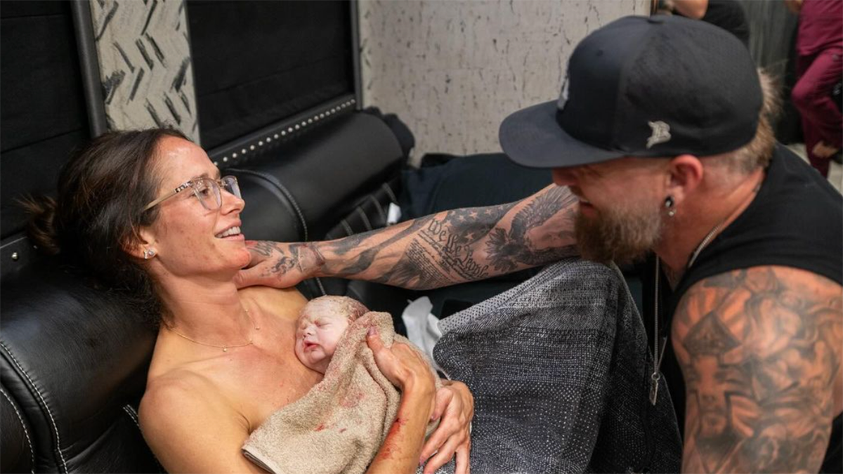 Brantley Gilbert stretches out his arm and touches his wife who swaddles her newborn baby in a towel