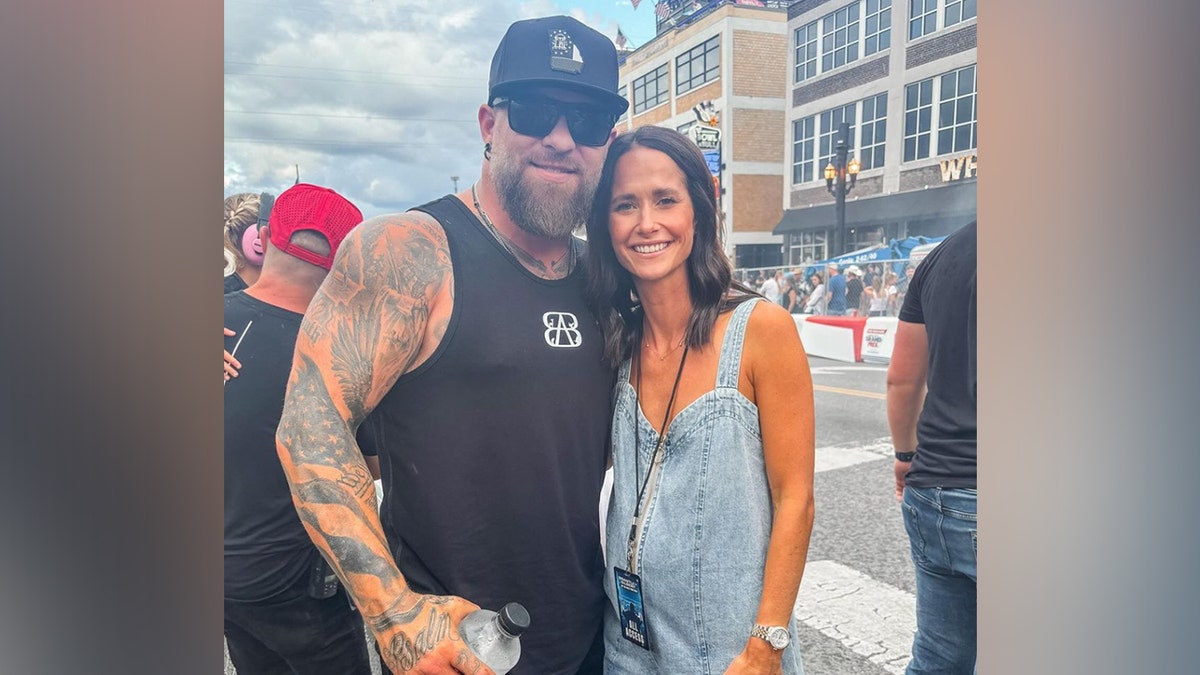 Brantley Gilbert in a black shirt with a hat and sunglasses smiles with his wife Amber in a denim romper