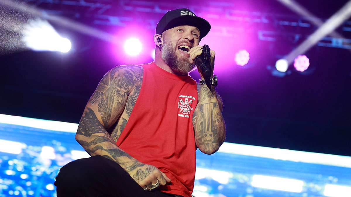 Brantley Gilbert sings on stage in a red cut-off shirt and black hat