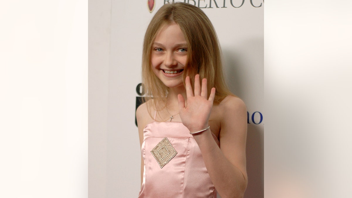 A young Dakota Fanning wears a light pink dress and waves on the carpet with braces