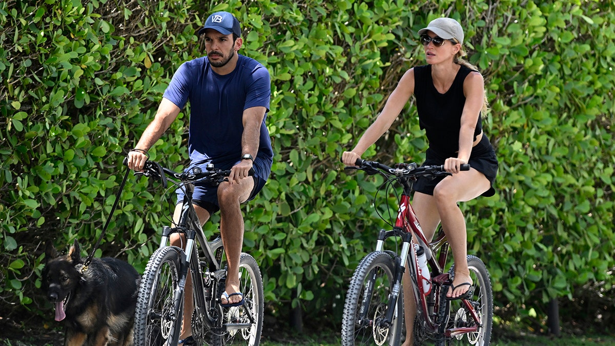 Joaquim Valente in a blue shirt and hat rides her bike next to Gisele Bündchen in a black outfit