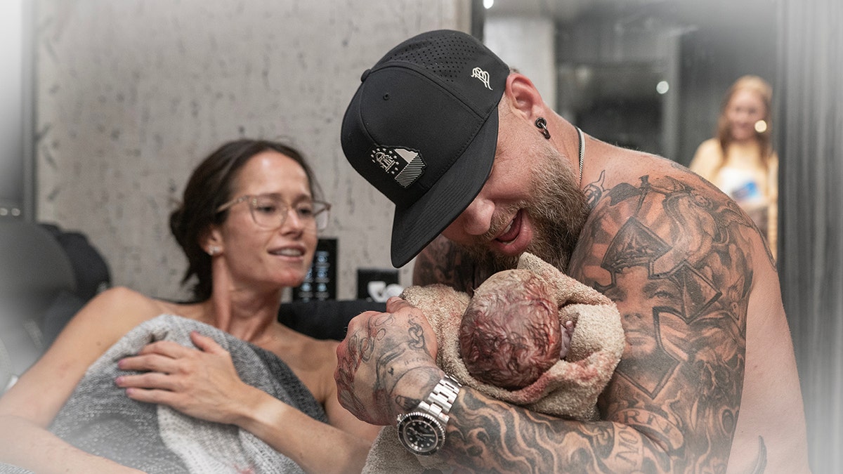 Brantley Gilbert holds his new baby wearing a black hat while his wife watches from under a blanket