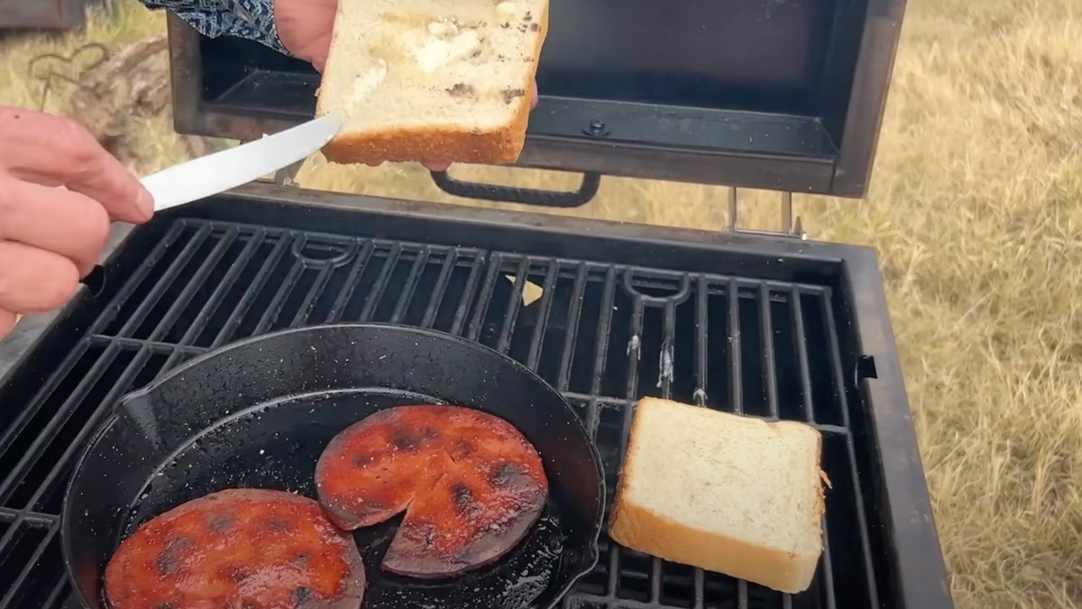 "Cowboy" Kent Rollins spreads butter on a piece of Texas toast with fried bologna cooking on a grill.