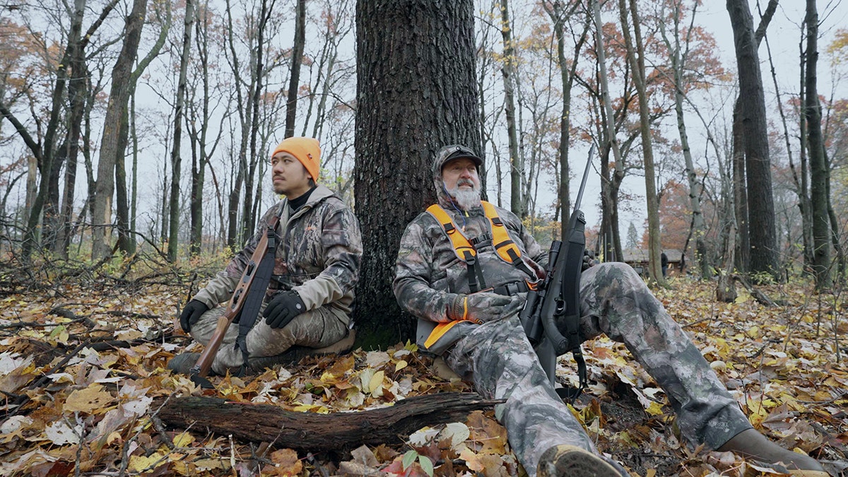 Two men sitting by a tree with hunting guns.