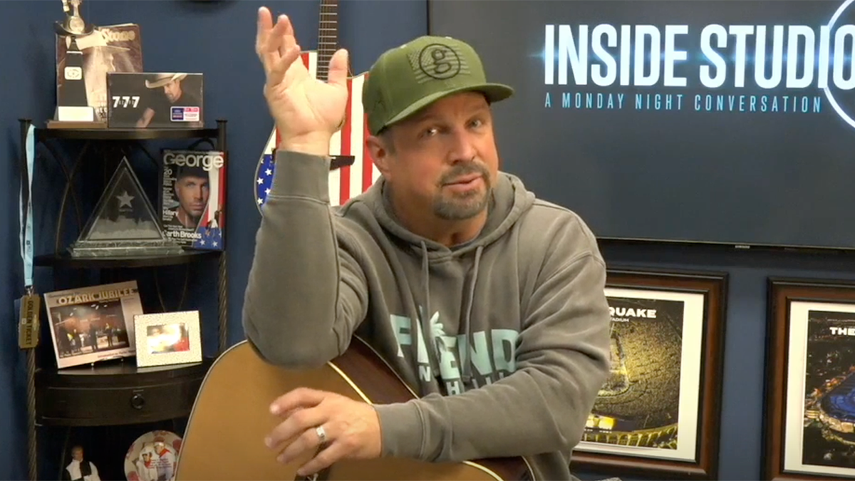 Garth Brooks con sudadera y gorra de béisbol verde levanta la mano mientras apoya la otra sobre su guitarra