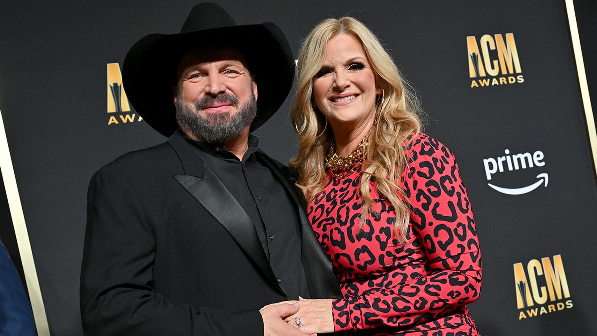 Garth Brooks in a black suit and black cowboy hat soft smiles with his wife Trisha Yearwood in a red cheetah dress