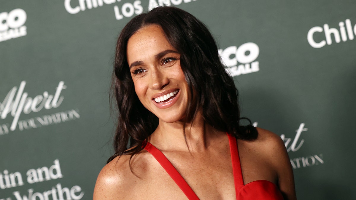 A close-up of Meghan Markle smiling wearing a plunging reddish dress.