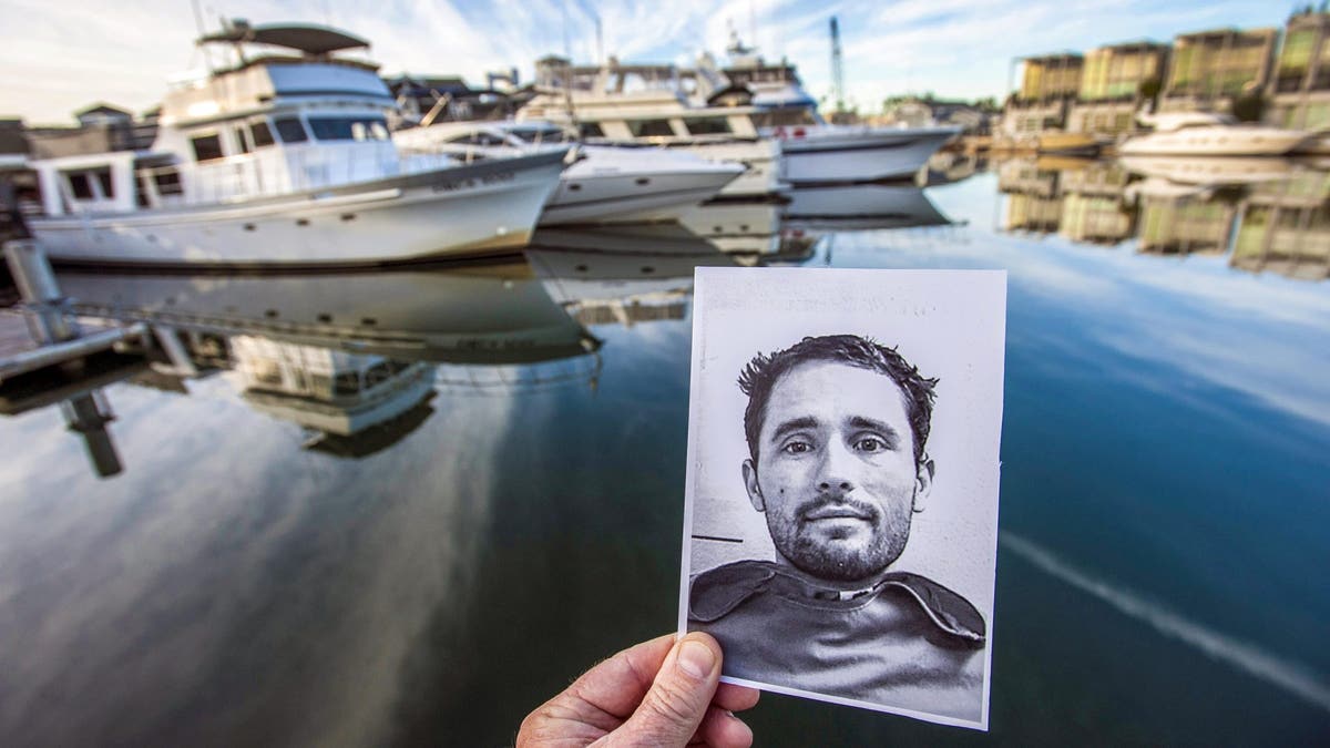 A person holds up a black and white mugshot of Skylar DeLeon pre-transition, with the Hawkses' 55-foot yacht anchored in the background