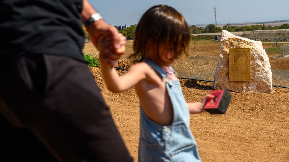 Una niña y su padre pasan junto a una roca y una fotografía del soldado asesinado Roni Eshel en un nuevo monumento a los soldados de vigilancia asesinados el 7 de octubre durante el ataque a la base de Nahal Oz el 4 de octubre de 2024, cerca de Nahal Oz, Israel. En la mañana del 7 de octubre, la base de Nahal Oz fue atacada por terroristas de Hamás, donde murieron 66 soldados, incluidas 15 mujeres soldado que operaban cámaras de vigilancia. Algunas de las mujeres soldado que no murieron fueron tomadas como rehenes el 7 de octubre de 2023.