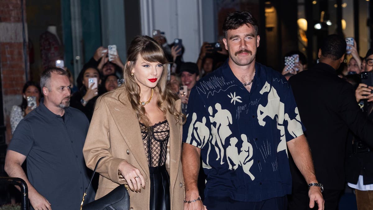 Taylor Swift in a brown jacket and black handbag holds Travis Kelce's hand in a dark blue shirt with figure/animal shapes drawn on it