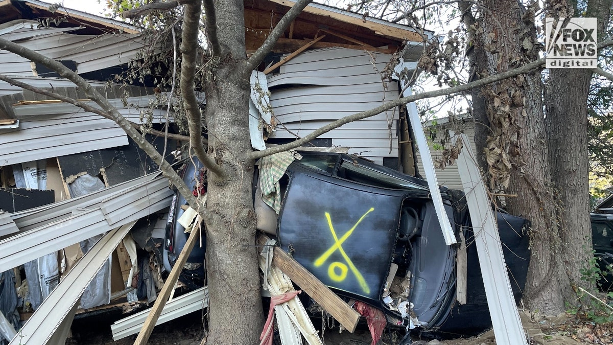 A car crushed between a house and a tree is colored yellow "X" spray-painted, meaning authorities found no one inside.