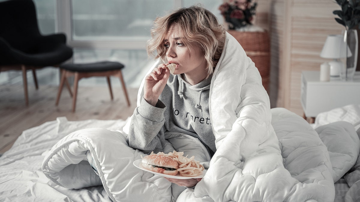 Mujer triste comiendo hamburguesa y patatas fritas en la cama