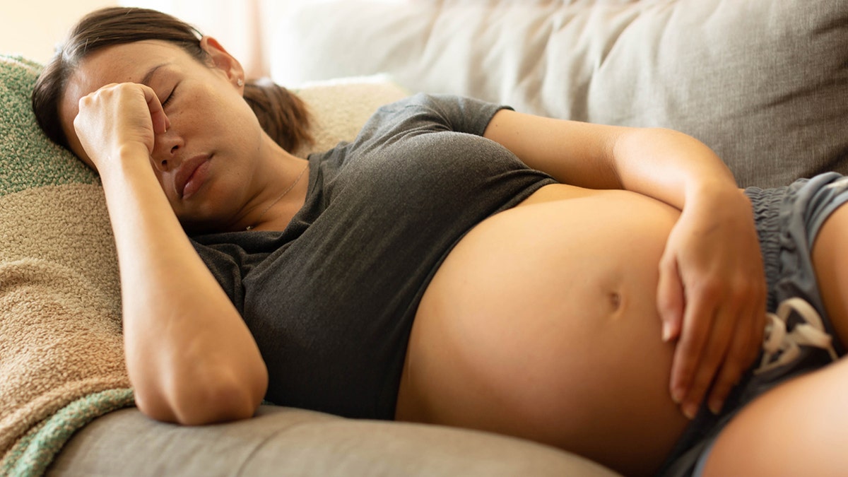 madre embarazada que intenta descansar en casa, tumbada en el sofá sujetándose la cabeza.