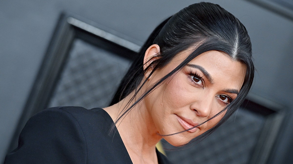 Kourtney Kardashian with a slicked back ponytail with pieces pulled out soft smiles on the carpet at the Grammys wearing a black jumpsuit