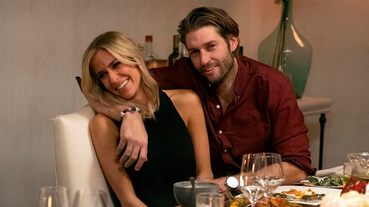 Kristin Cavallari de blusa preta se inclina enquanto se senta à mesa com Jay Cutler, que está vestindo uma camisa marrom e tem o braço em volta de Kristin