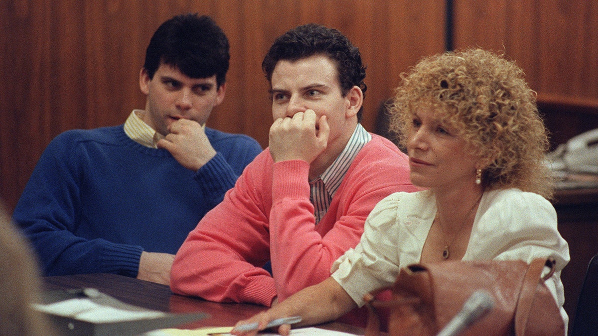 Lyle Menendez in a blue sweater and Kyle Menendez in a coral sweater sit with attorney Leslie Abramson, both with their hands on their mouths and chins