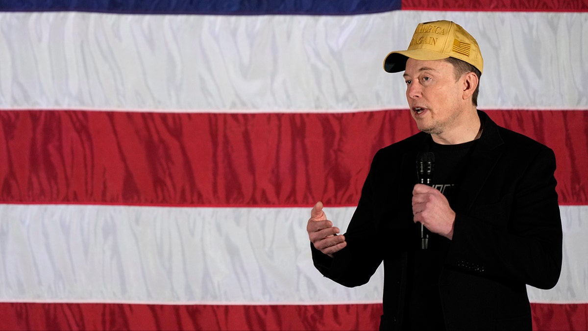 Elon Musk in yellow hat with US flag behind him