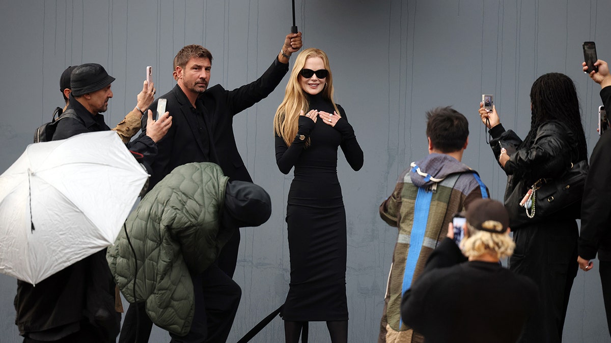 Nicole Kidman in a long black dress holds her hands to her chest as photographer crouch down to take snap a picture