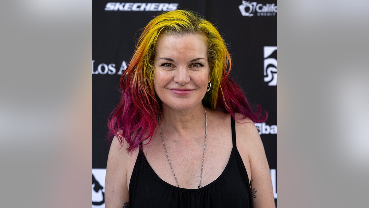 Pauley Perrette con un top negro y el pelo ombré multicolor amarillo y rosa oscuro sonríe suavemente en la alfombra