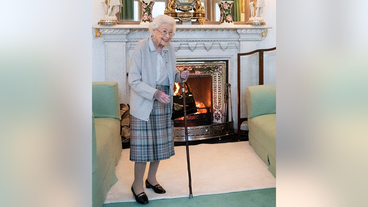 A frail Queen Elizabeth in a plaid skirt and light grey sweater smiles while holding a cane and wearing glasses