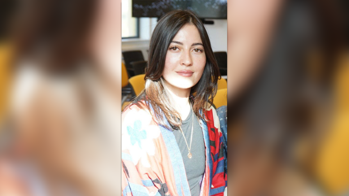 Sabina Rosas sits in a classroom with her hair down and sweats in a colorful sweater over a T-shirt