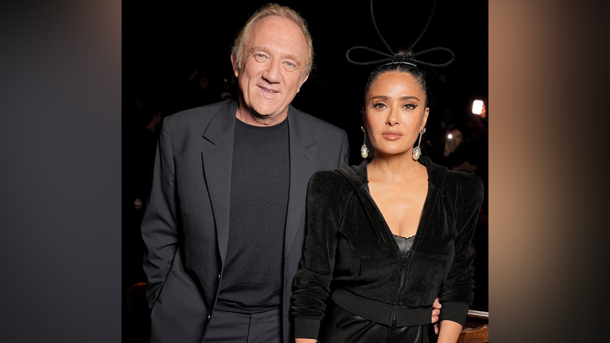 François-Henri Pinault, in a gray suit and black shirt, smiles next to his wife Salma Hayek in a black dress and velor jacket at the Balenciaga RTW Spring show