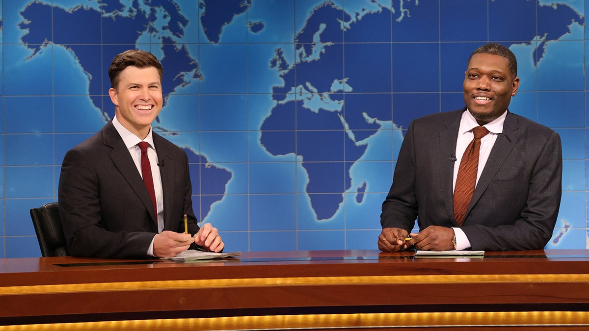 Colin Jost and Michael Che both laugh behind the Weekend Update desk in dark suits with red ties