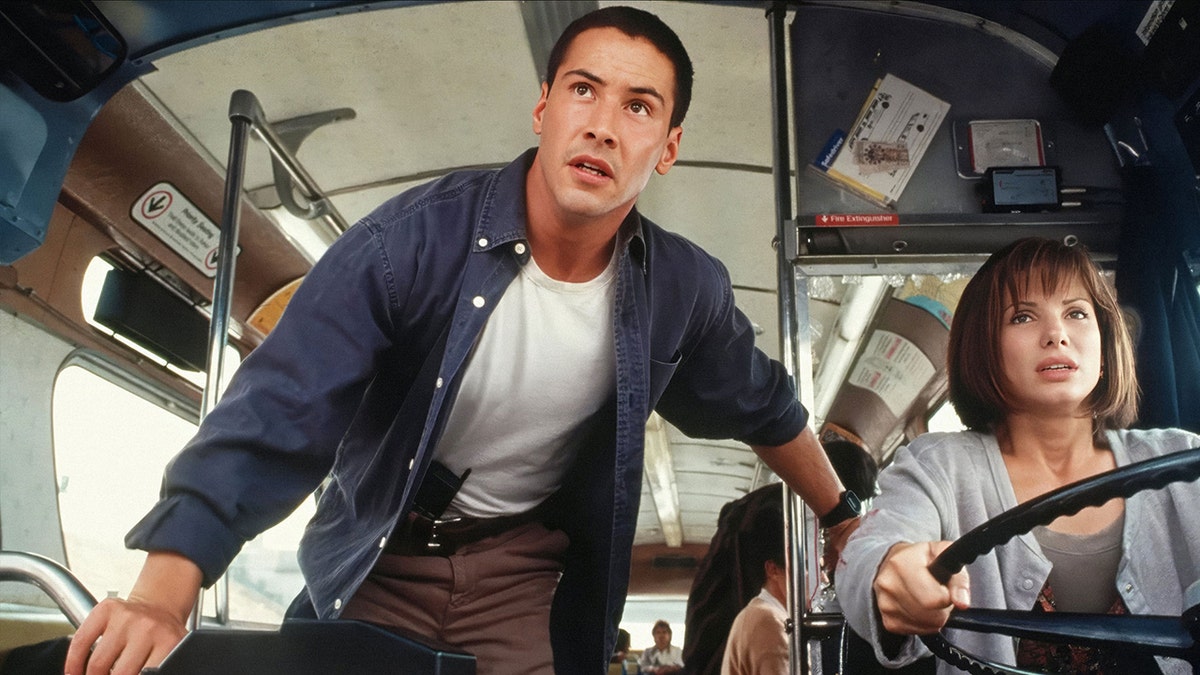 Keanu Reeves in a white t-shirt and blue jean shirt looks up while Sandra Bullock in a grey sweater holds the wheel of a vehicle