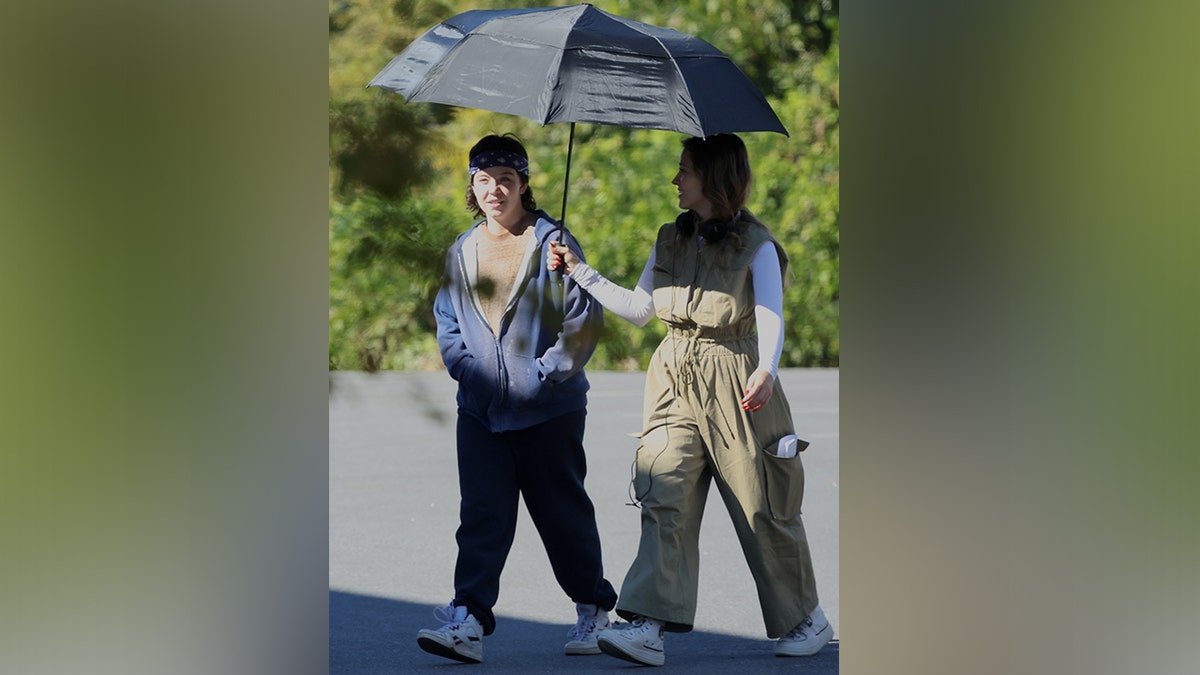 Sydney Sweeney dengan celana olahraga berjalan di lokasi syuting dengan seseorang memegang payung hitam di atasnya