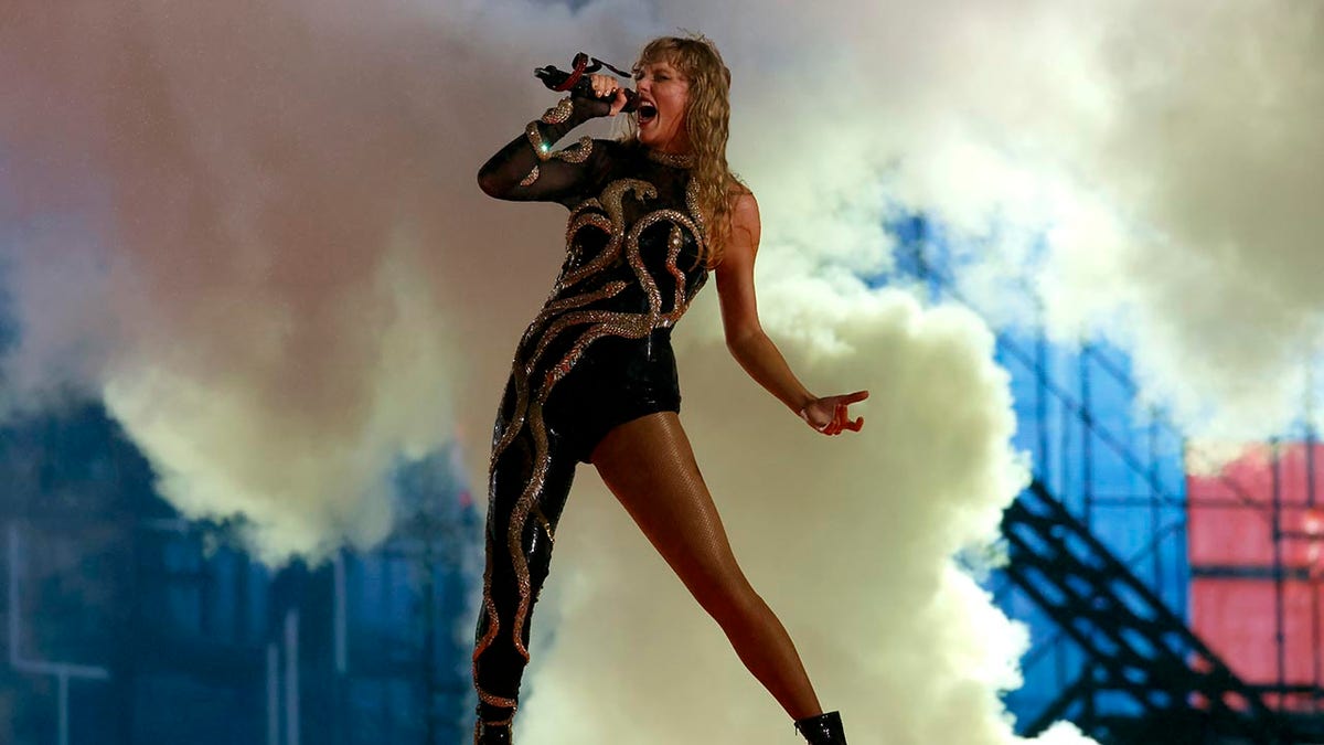 Taylor Swift con su nuevo conjunto de 'Reputation', un mono con una sola pierna completamente cubierta, canta dramáticamente ante el micrófono.