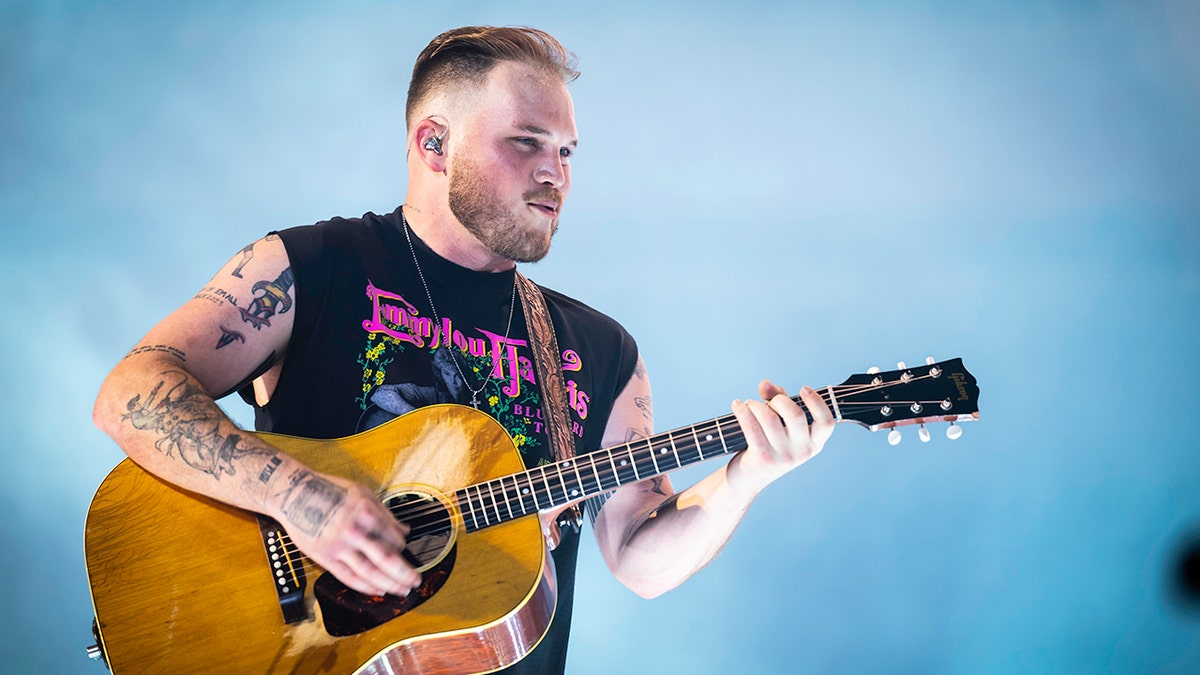 Zach Bryan in a cut-off black tank top strums his guitar on stage