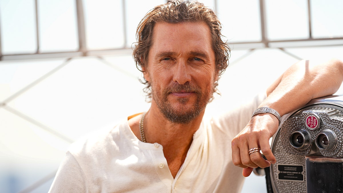 Matthew McConaughey leans his arm against a telescope/magnifier at the top of the Empire State Building, wearing a loose white shirt