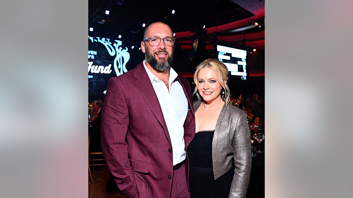 Mark Wilkerson in a cranberry red suit smiles next to wife Melissa Joan Hart in a black dress and metallic jacket