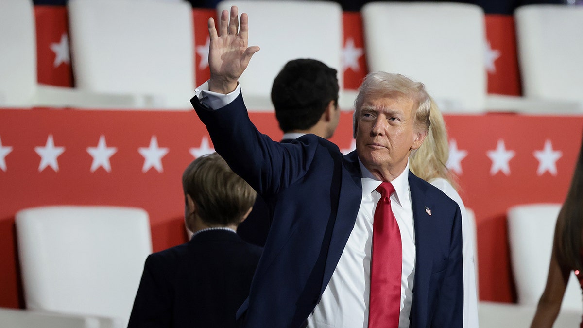 Donald Trump con traje oscuro y corbata roja saluda a la multitud y mira a su derecha