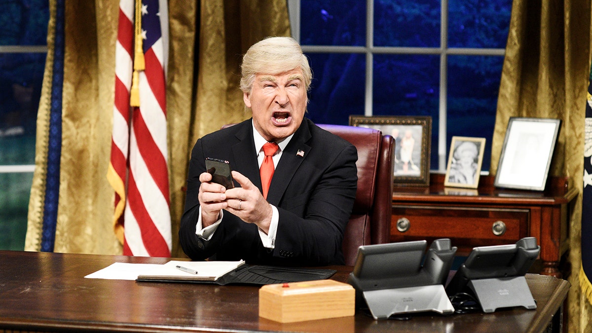 Alec Baldwin vestido como el Presidente Donald Trump  con traje negro y corbata roja sostiene un teléfono en sus manos y parece disgustado