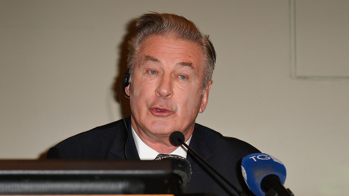 Alec Baldwin in a black suit speaks into a microphone during a press conference at the Torino Film Festival.