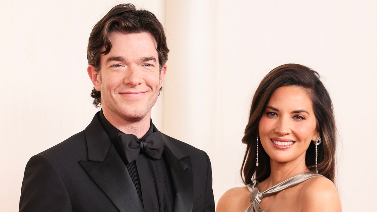 John Mulaney in an all-black tuexedo soft smiles on the Oscars carpet with Olivia Munn in a halter silver dress