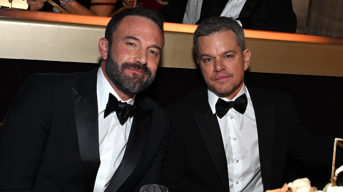 Ben Affleck and Matt Damon both in tuxedos soft smile at a table at the Golden Globes