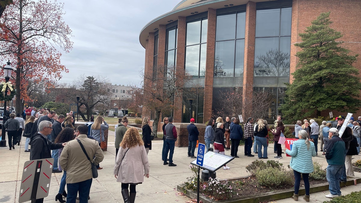 People in jackets stand in line while others join them outside a round brick building with large windows.