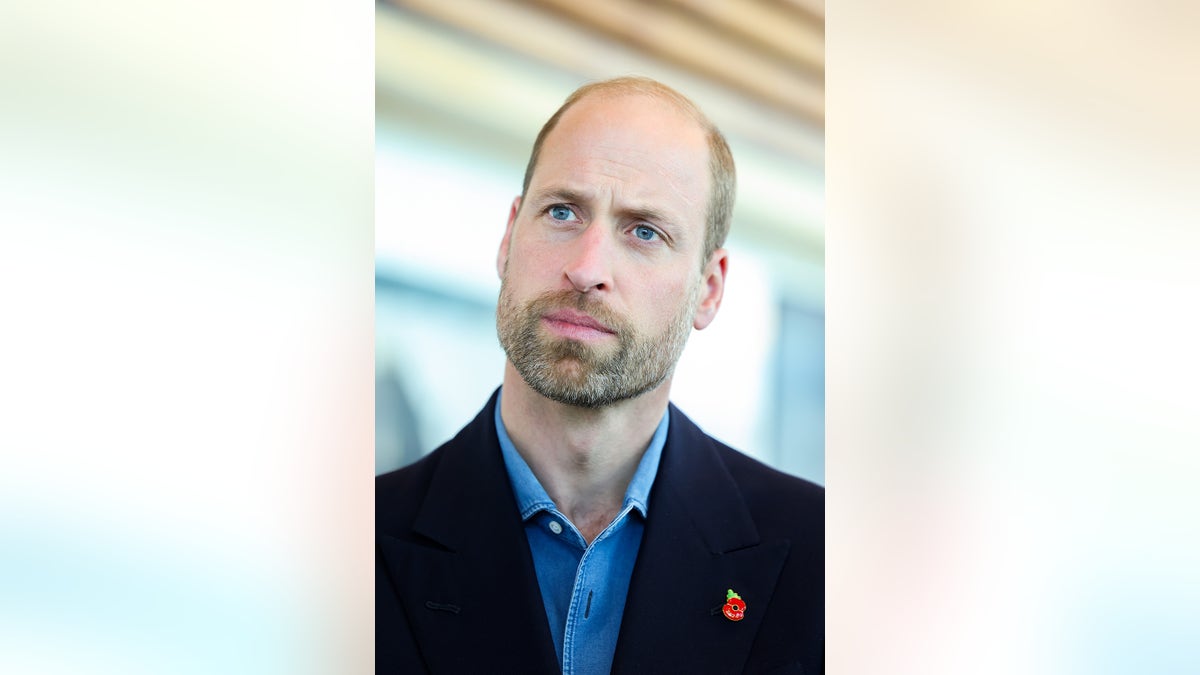 Close-up of Prince William looking serious wearing a dark jacket and blue shirt with a red pin.