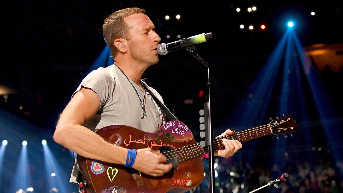 A picture of Chris Martin taken from the side, as he strums the guitar on stage wearing a grey shirt