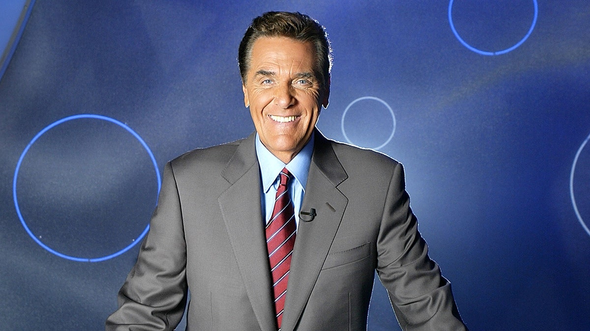 Chuck Woolery successful  a grey suit, bluish  garment  and reddish  striped necktie  smiles for a photo